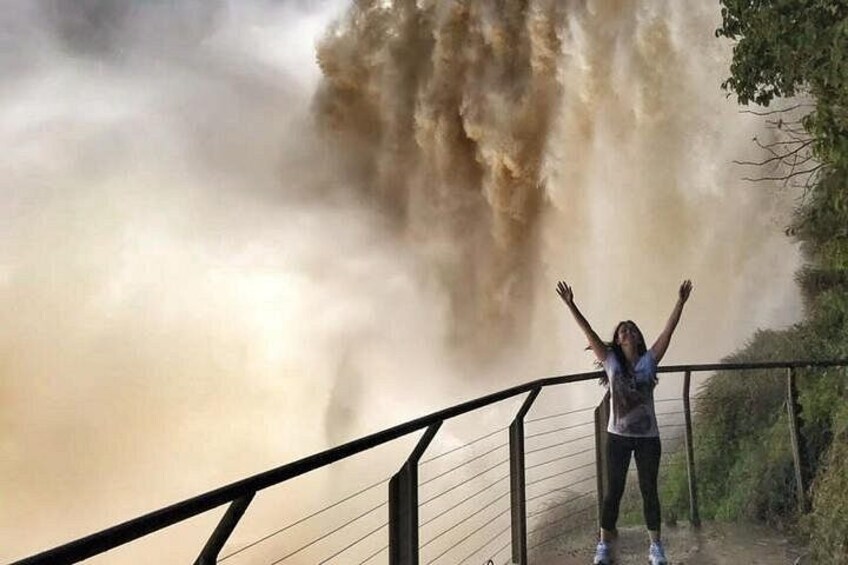 A person standing on a bridge railing with arms raised, overlooking a large body of water with a turbulent, brownish surface. 1