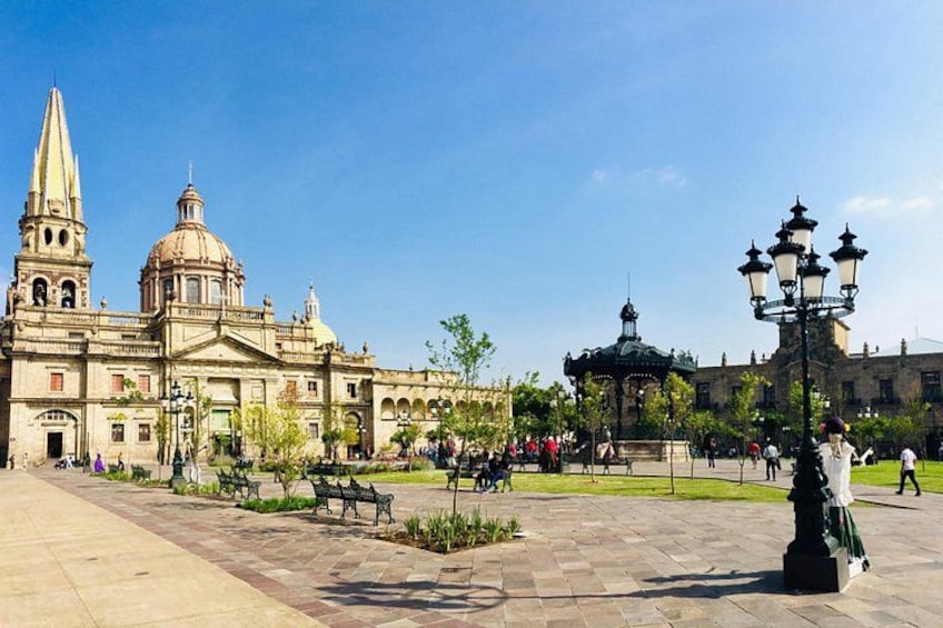 Un edificio histórico con cúpula y aguja, un pabellón, y una plaza empedrada con bancos y gente. 2