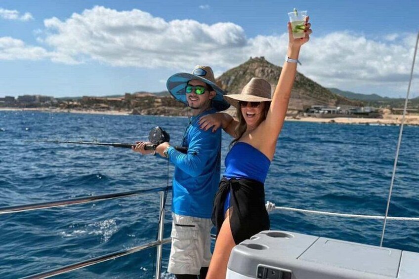 Two people on a boat, one holding a fishing rod, with a beach and mountains in the background. 2