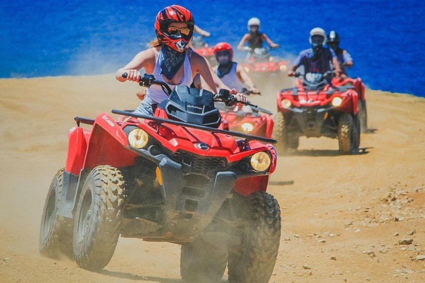 People riding red ATVs on a sandy beach with a body of water in the background. 2
