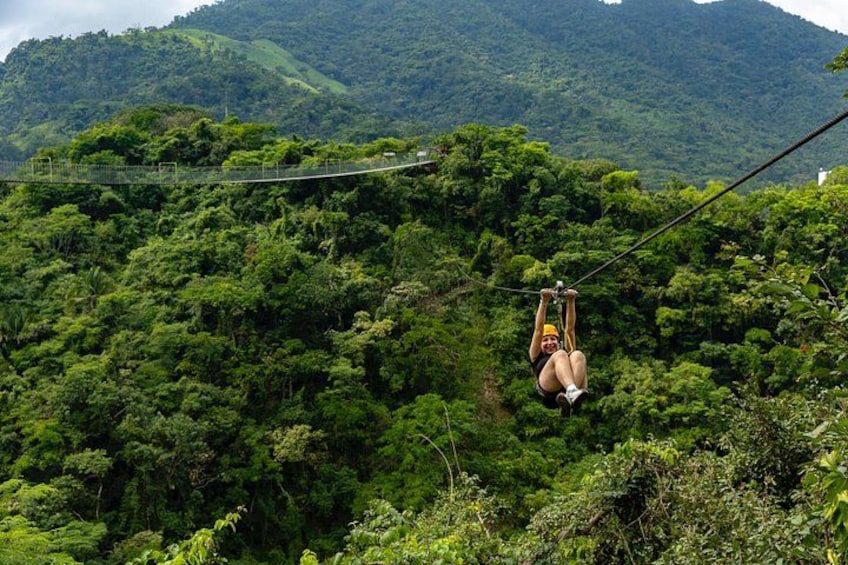 A person suspended on a zip line over a dense forest with a mountain in the background. 1