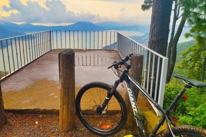 En mountainbike parkeret på en våd træplatform med gelænder, med udsigt over vand og bjerge. 2