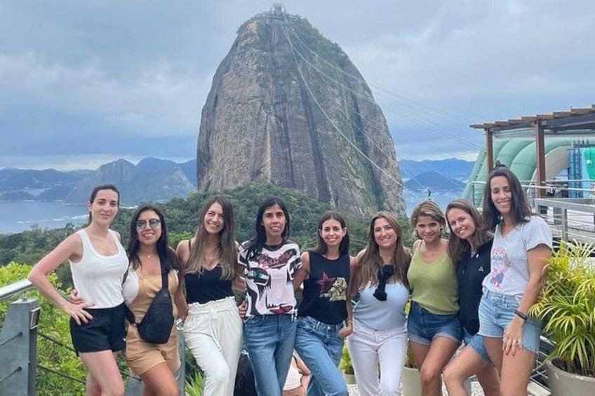 A group of people posing in front of a large rock formation with a view of the ocean and mountains in the background. 2