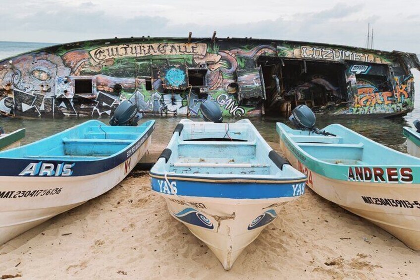 Three small boats named ARIS, YAS, and ANDRES on a sandy beach with a large, graffiti-covered ship in the background. 2