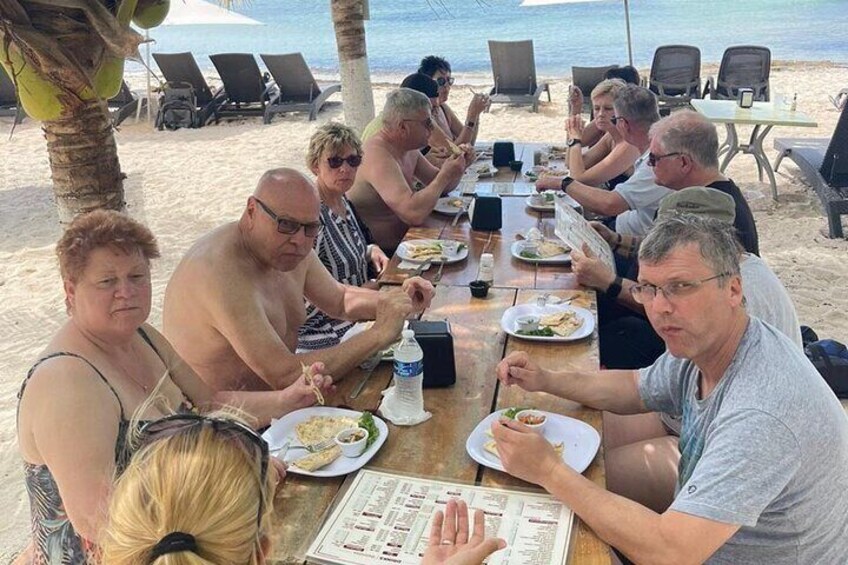A group of people dining at a beachside table with plates of food, palm trees, and the ocean in the background. 4