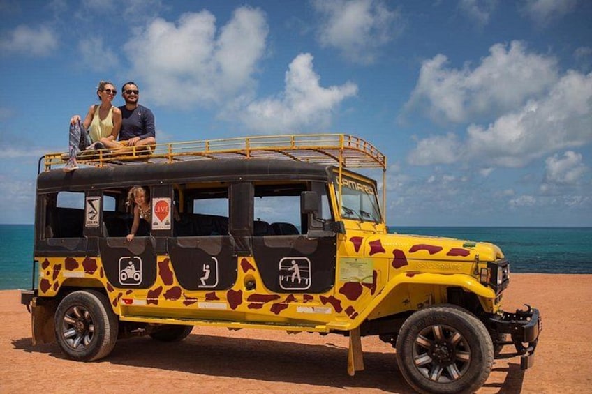 A yellow safari vehicle with a person and a child on top, parked on a sandy beach with the ocean in the background. 1