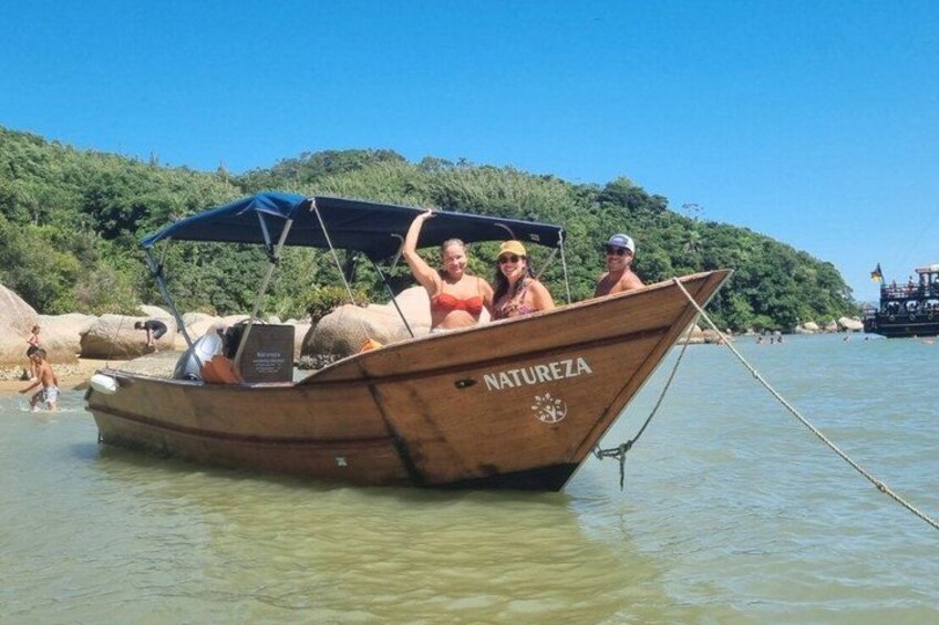 A wooden boat named 'NATUREZA' with a canopy, docked at a beach with a forested hill in the background. 3