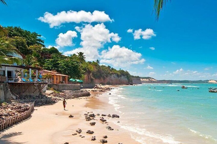 Una playa con agua turquesa cristalina, una orilla de arena y algunas embarcaciones. Se ven palmeras y un edificio con balcón. 2