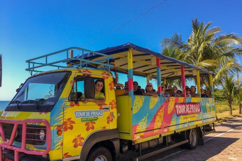 A colorful bus with 'TOUR DE PRAIAS' written on it, parked on a road with palm trees in the background. 4