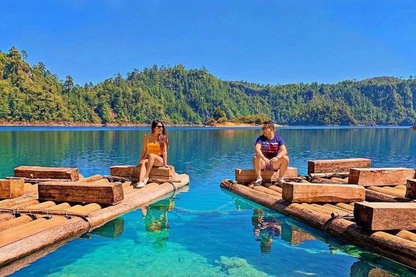 Two people sitting on wooden platforms in a clear blue lake surrounded by forest. 1