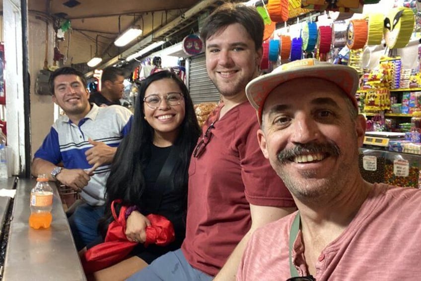 Un groupe de personnes se trouve sur un marché où sont exposés des textiles aux couleurs vives et divers produits. 2