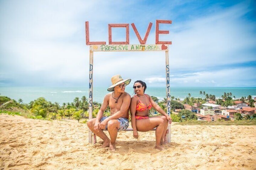 Deux personnes sont assises sous un panneau représentant le mot « AMOUR », sur lequel on peut lire « PROTÉGEONS LA NATURE », sur une plage d’où l’on aperçoit l’océan et une ville en arrière-plan. 3