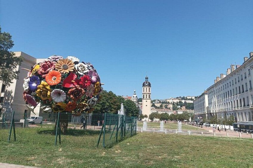 En stor blomsterskulptur i en offentlig plads med et historisk tårn i baggrunden. 4