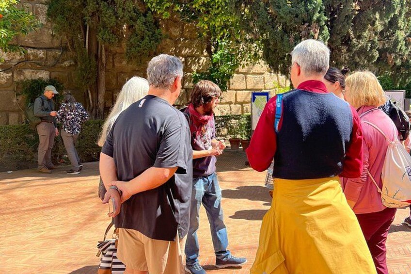Un grupo de personas de pie al aire libre, posiblemente en un parque o sitio histórico, con árboles y muros de piedra al fondo. 2