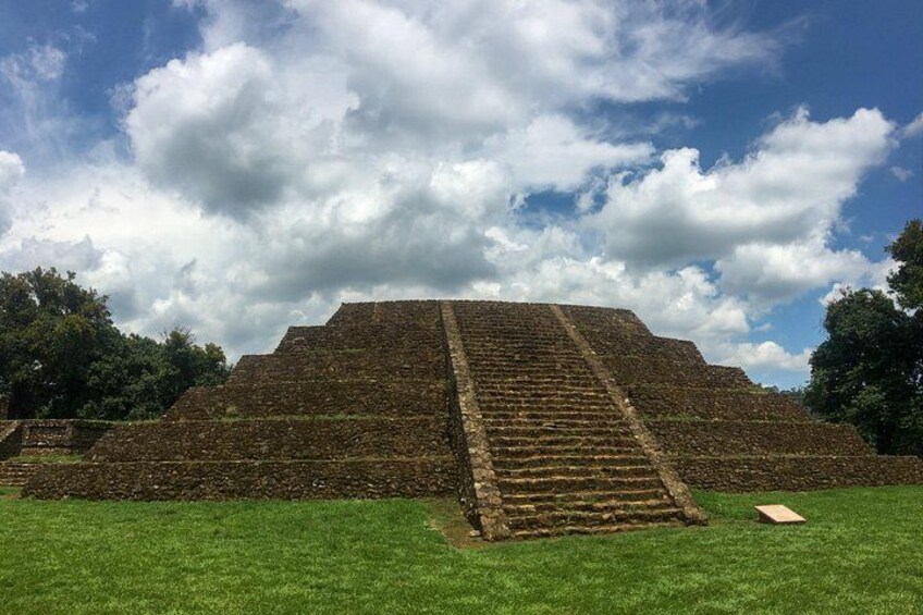 A large pyramid with a staircase leading up to the top, surrounded by green grass and trees under a partly cloudy sky. 2