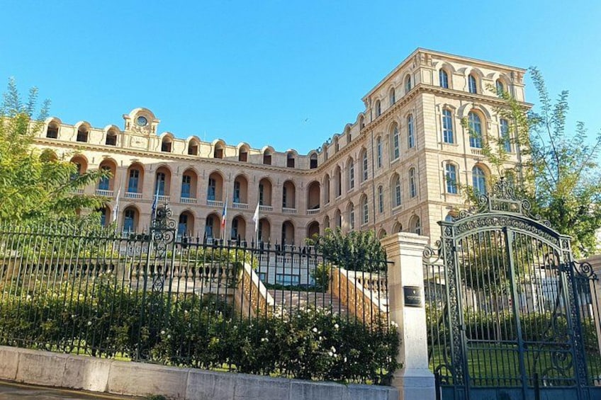 Un grand bâtiment historique doté d’arcades et d’une tour horlogère centrale, entouré d’un jardin bien entretenu et accessible par une entrée fermée par un portail. 2