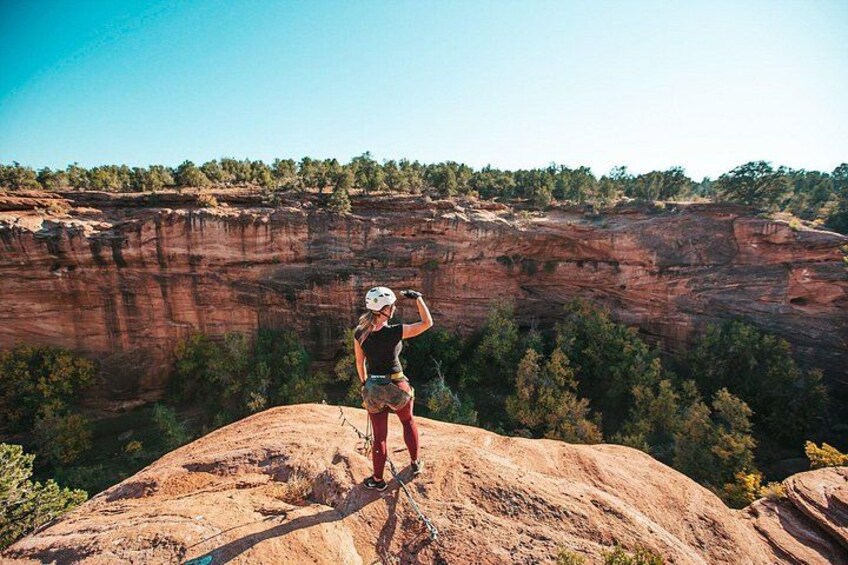 A person standing on a rock with a helmet, overlooking a canyon with red rock formations and trees. 3