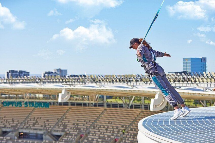 Una persona realiza una acrobacia aérea en una plataforma circular con una cuerda, con un estadio y edificios al fondo. 4