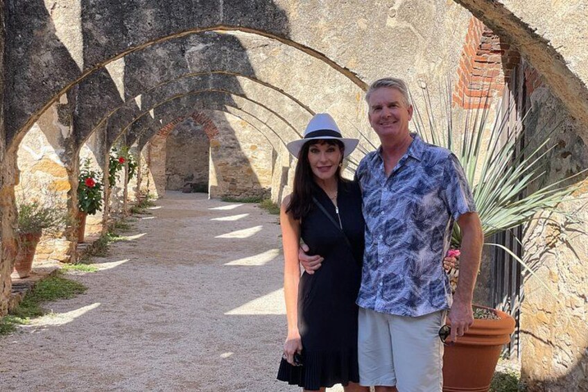 Un viale in pietra con archi, una donna in abito e cappello neri e un uomo con una camicia fantasia. 2