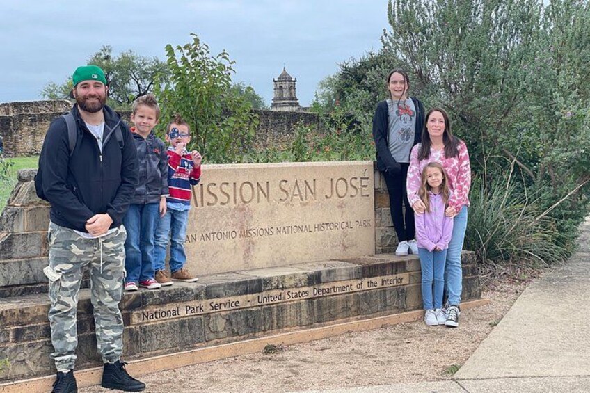 Un gruppo di persone in piedi di fronte a un cartello con su scritto 'MISSION SAN JOSÉ', con un edificio storico sullo sfondo. 5