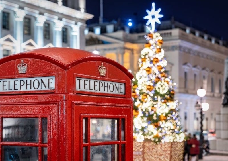 Una cabina telefónica roja con letreros que indican 'TELEPHONE', un árbol de Navidad decorado y un edificio al fondo. 1