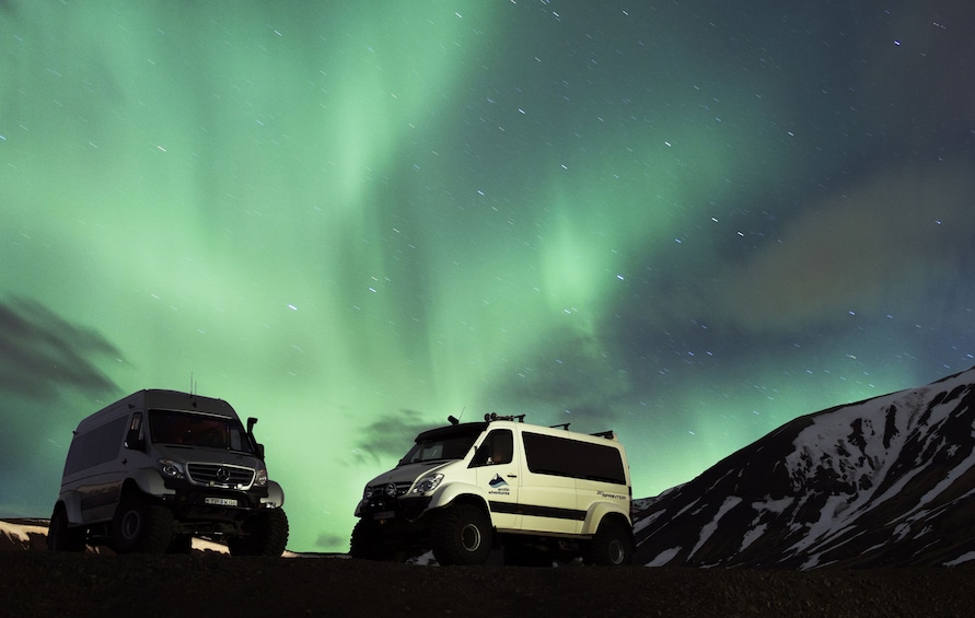 Two white vans parked on a snowy mountain under a green aurora. 1