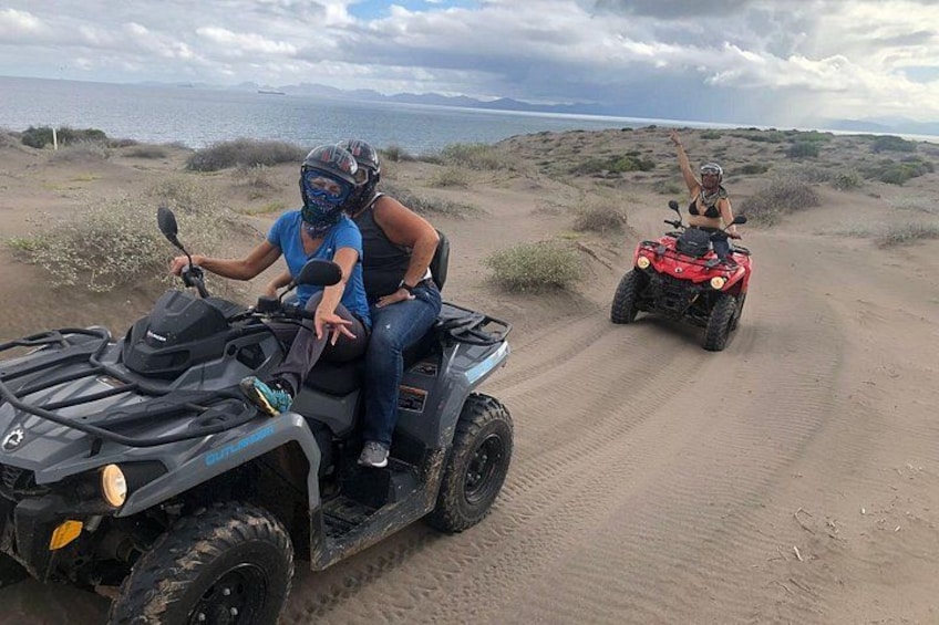 Two people on ATVs on a sandy terrain with sparse vegetation and a distant body of water. 1