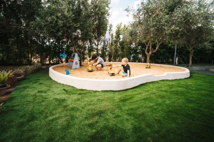 Ein runder Sandkasten mit spielenden Kindern, umgeben von einer grünen Rasenfläche und Bäumen. 3