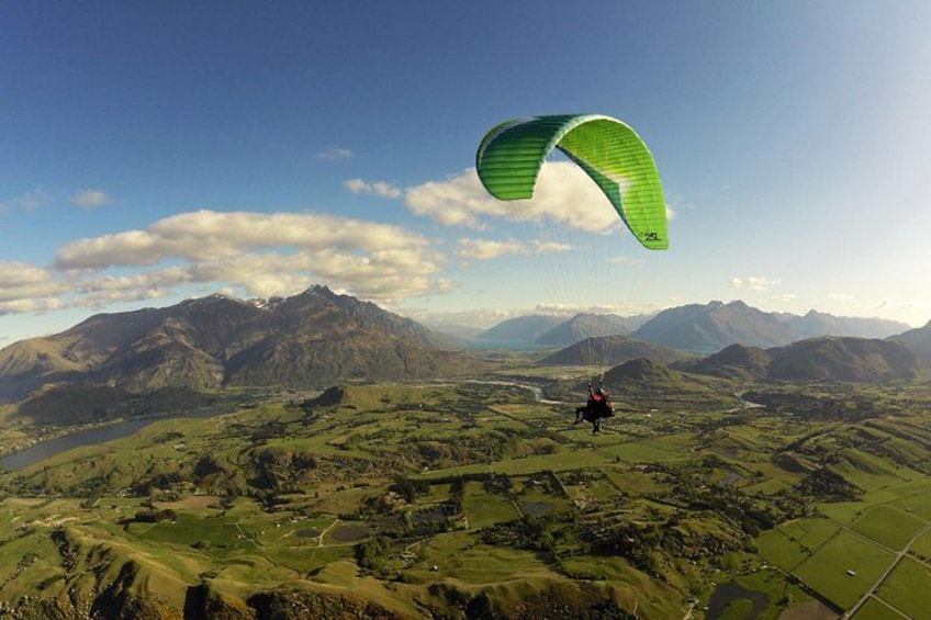 Una persona practica parapente sobre un paisaje montañoso con campos verdes y montañas al fondo. 1
