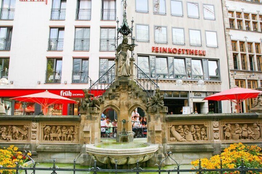 Ein Brunnen mit Skulpturen vor einem Gebäude, dessen Schild "FEINKOSTHEKE" zeigt. 4