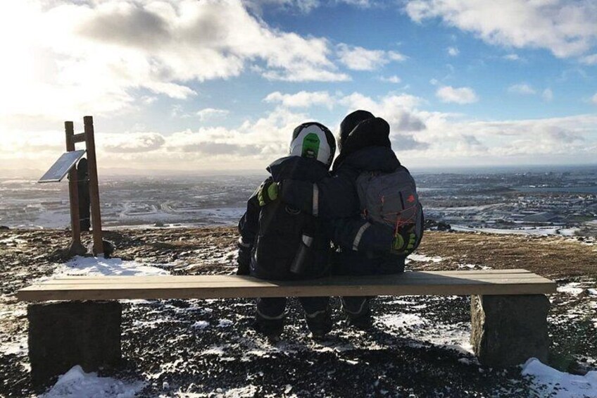 Två personer sitter på en bänk med ryggsäckar och blickar ut över ett snöigt landskap och en avlägsen stad. 3