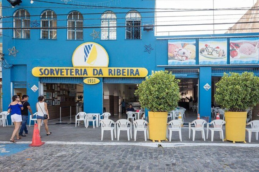 A blue building with the sign 'Sorveteria da Ribeira 1931'. There are outdoor tables and chairs, and two large yellow planters. 3