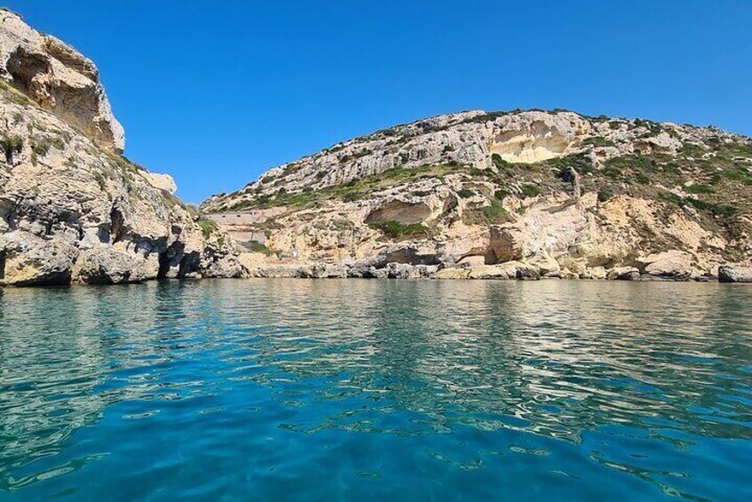 A coastal landscape with clear blue waters, rocky cliffs, and a hilly terrain. 1