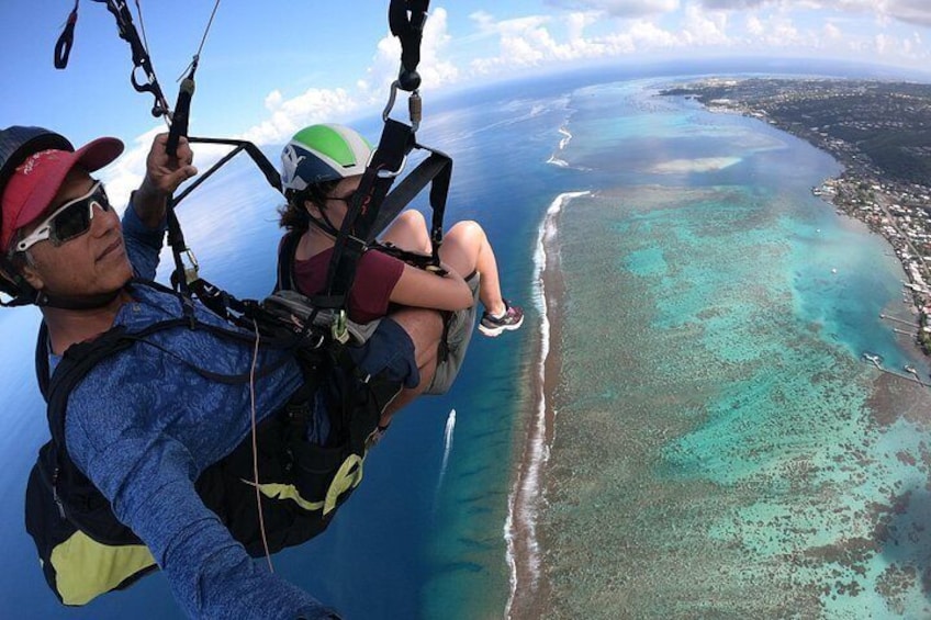 A person in a blue jacket and red cap, holding a camera, with a child in a green helmet and red shirt, both wearing harnesses, flying over a coastal area with clear turquoise waters and a sandy beach. 1