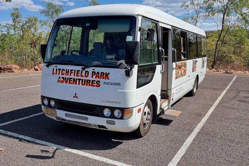 Een witte bus met 'Litchfield Park Adventures' op de voorkant, geparkeerd aan een weg met bomen en rotsen op de achtergrond. 4