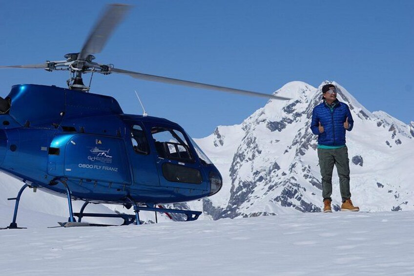 A blue helicopter with a person standing beside it on a snowy mountain. 1