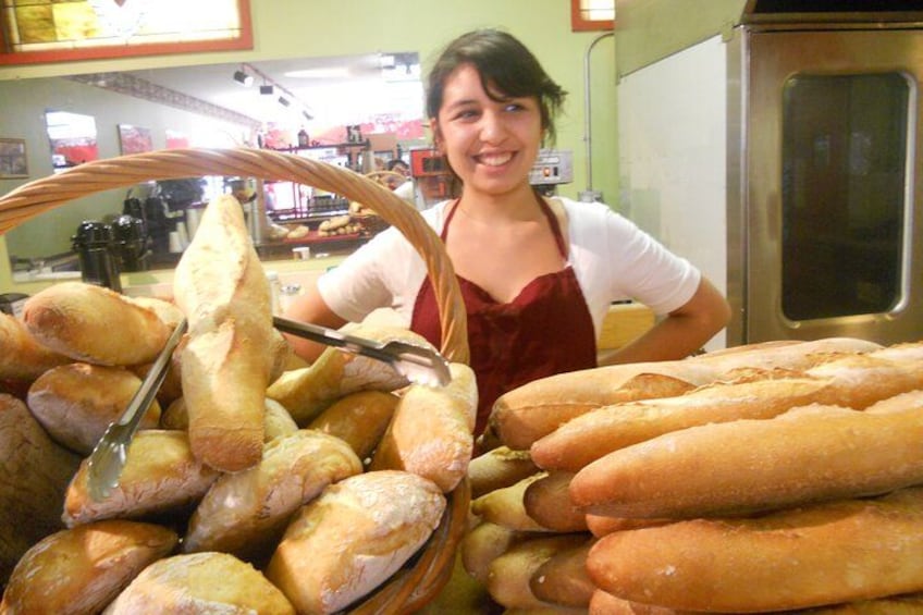 En person holder et baguettebrød, med en kurv fyldt med baguettebrød i en bagerbutik. 5