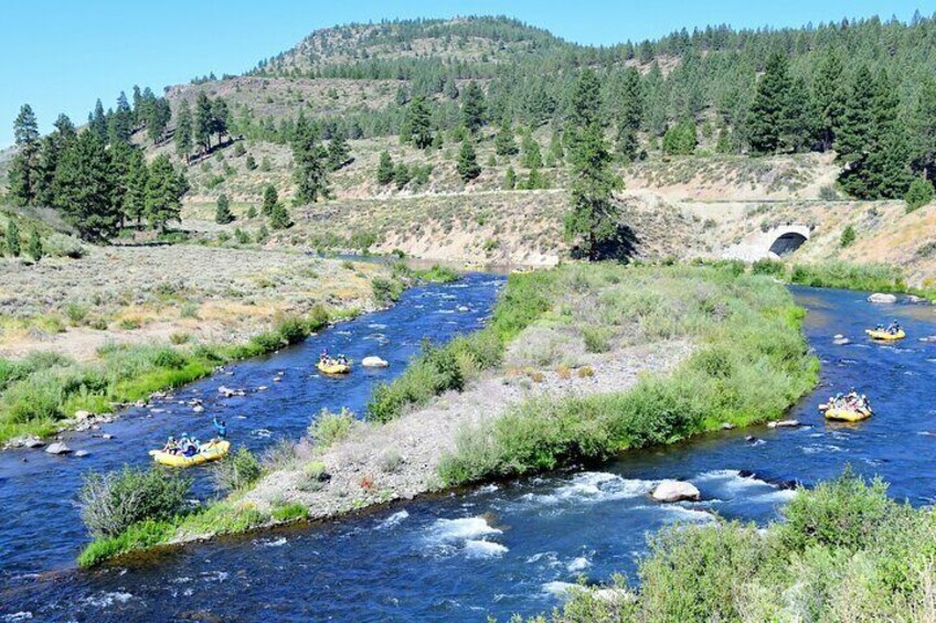 A river with multiple yellow rafts navigating through it, surrounded by greenery and a bridge in the background. 2