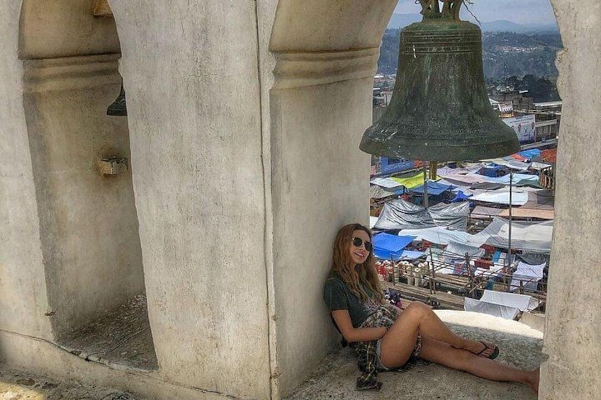 A person sitting in a stone structure with a large bell in the background. 5