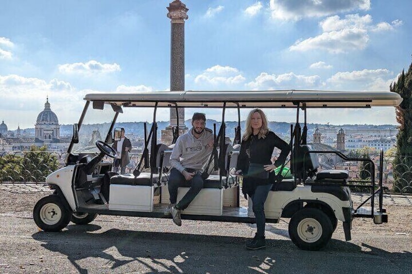 A golf cart with two people, one driving and one standing, in front of a cityscape with a prominent tower and dome. 2