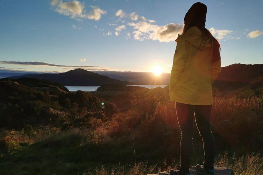 A person standing on a grassy hill overlooking a lake with mountains in the background during sunset. 2
