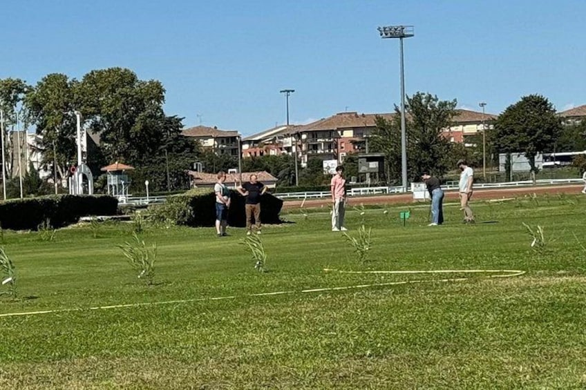 A grassy field with people walking and standing, a track, and buildings in the background. 5