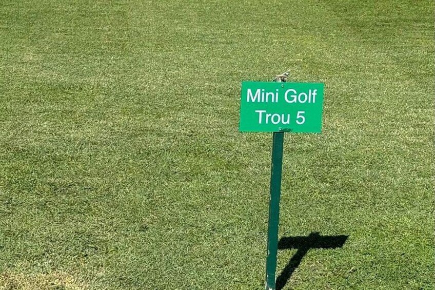 A green sign with white text reading 'Mini Golf Trou 5' on a grassy field. 3