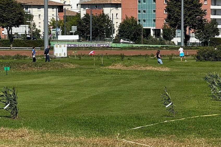 A grassy field with people playing, a sign reading 'TOULOUSE', and buildings in the background. 4