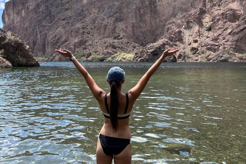 En person i bikini som står i vattnet med armarna lyfta och vänder sig mot en klippig brant. 2