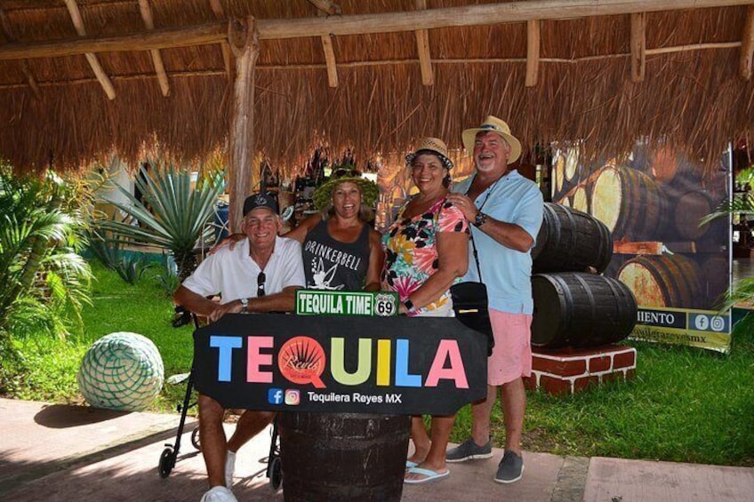 A group of people posing in front of a sign that reads 'TEQUILA TIME 69' with a backdrop of barrels and a thatched roof structure. 2