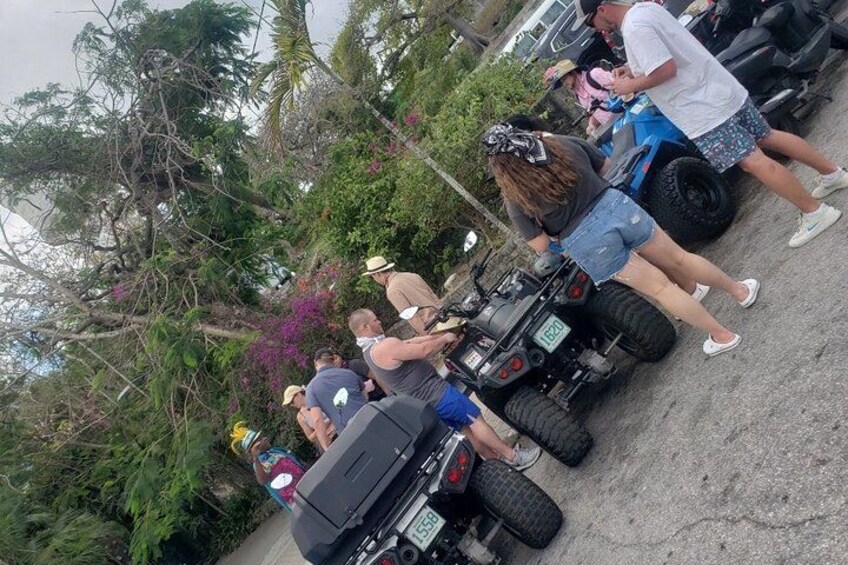 People around ATVs in a parking area with trees and purple flowers in the background. 5