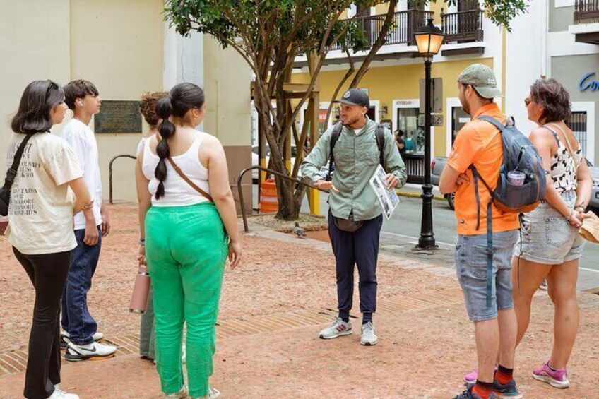 A group of people standing on a brick-paved area, some with backpacks, in an outdoor setting with buildings and a tree in the background. 1