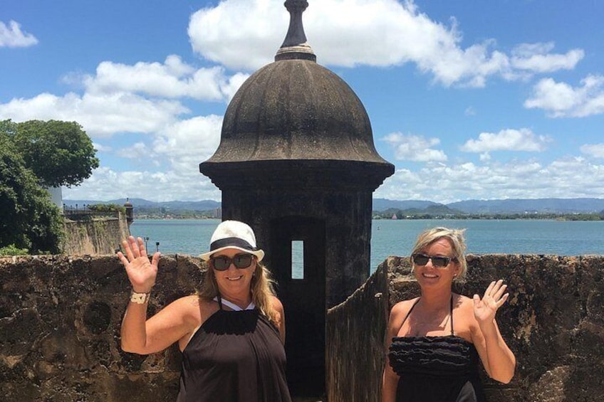 Two people standing in front of a historical stone structure with a body of water and mountains in the background. 5