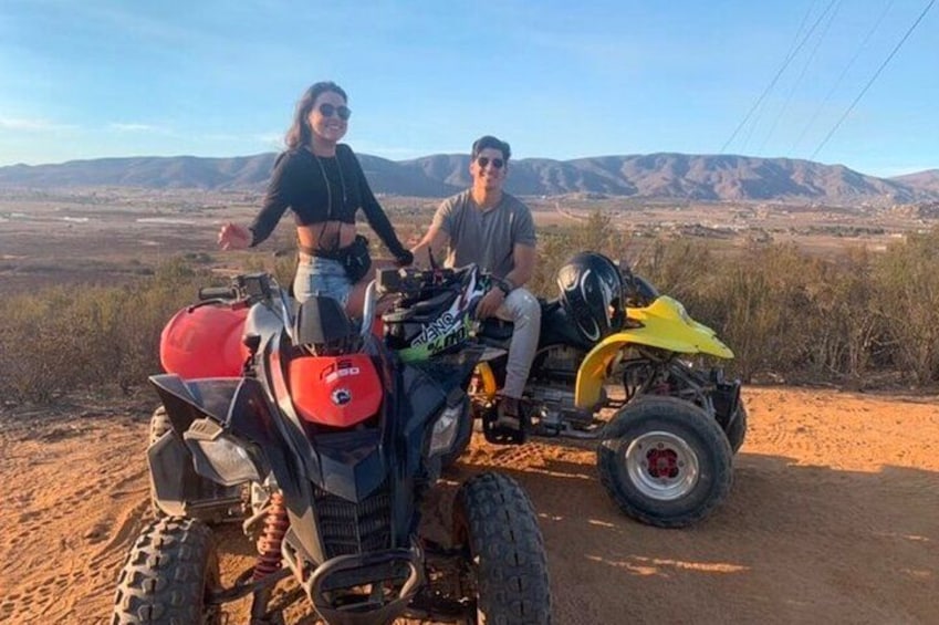 Two people on a yellow ATV in a desert landscape with mountains in the background. 3